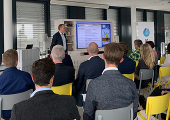 Eine Person präsentiert einer Gruppe von Menschen etwas auf einem großen Screen, sie sitzen und hören zu